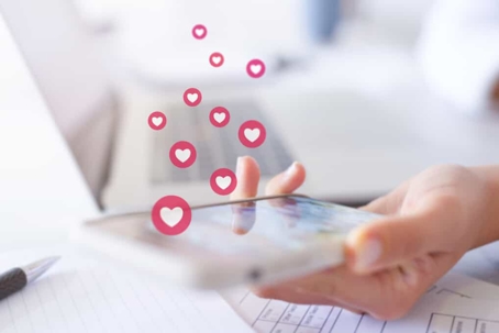 Hand holds a smartphone over a laptop in a brightly lit room with social media heart "likes" coming up in the frame from the phone